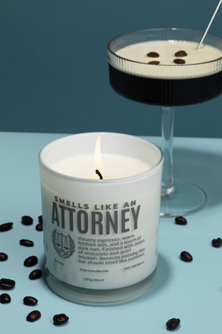 Candle labeled 'Smells Like an Attorney' next to a cocktail with chocolate shavings on a blue surface.