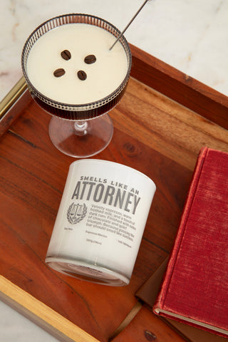 Cocktail with espresso beans  in a glass on a wooden tray next to a candle labeled 'Smells Like an Attorney'.