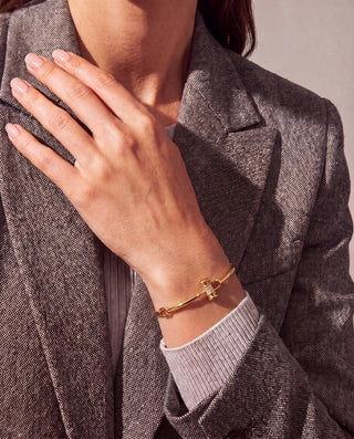 A close-up of a person wearing a delicate gold bracelet with a small central charm, styled on their wrist with a gray blazer and ribbed shirt visible.