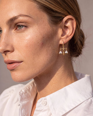 A close-up of a woman wearing a delicate gold dangle earring shaped like a balance scale with two small white triangular elements, paired with a white collared shirt.