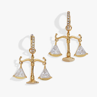 A pair of gold-tone balance scale earrings with clear triangular gemstones on the scales and small clear stones along the hooks, shown on a light gray background.